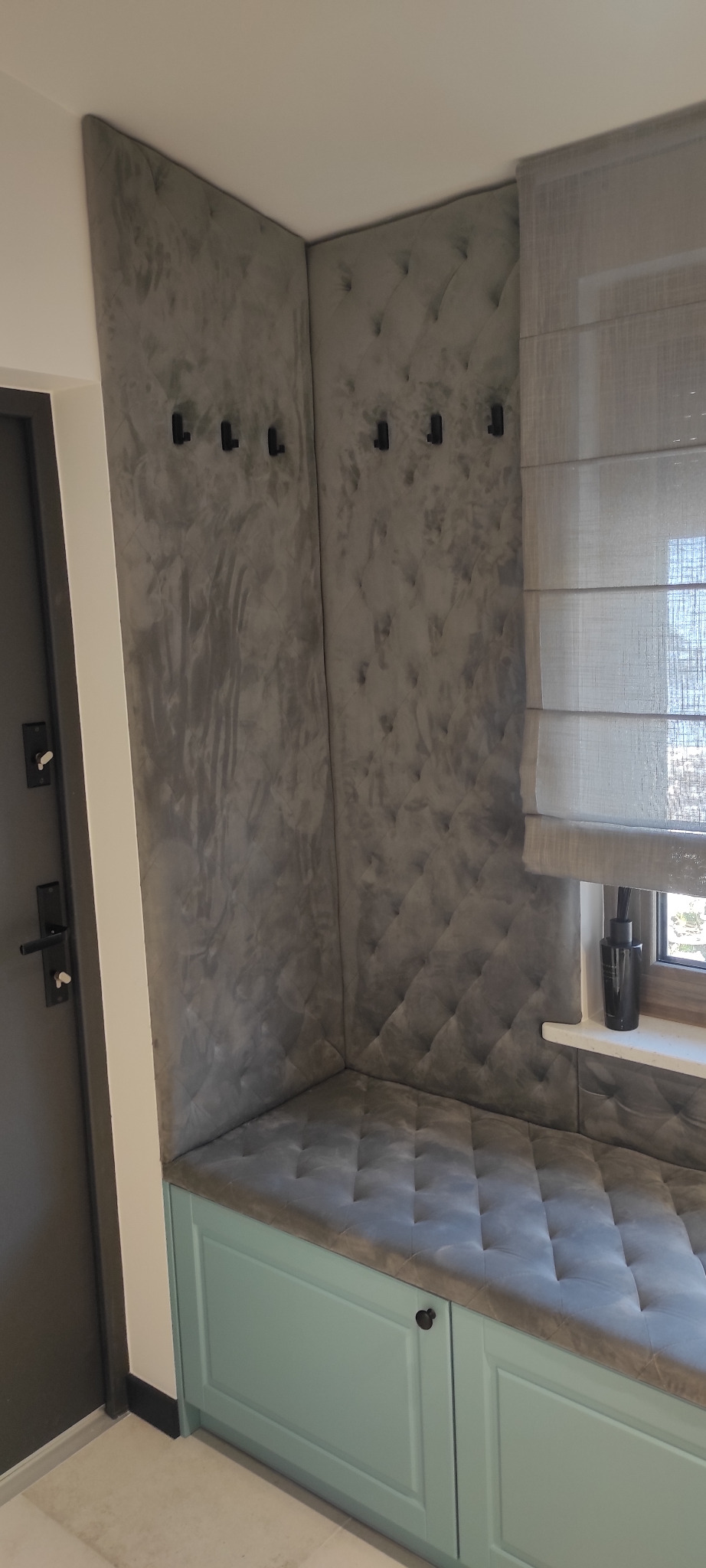 Wnęka z siedziskiem i tapicerowanymi ścianami w odcieniach szarości, z haczykami na ubrania i roletą rzymską w oknie.