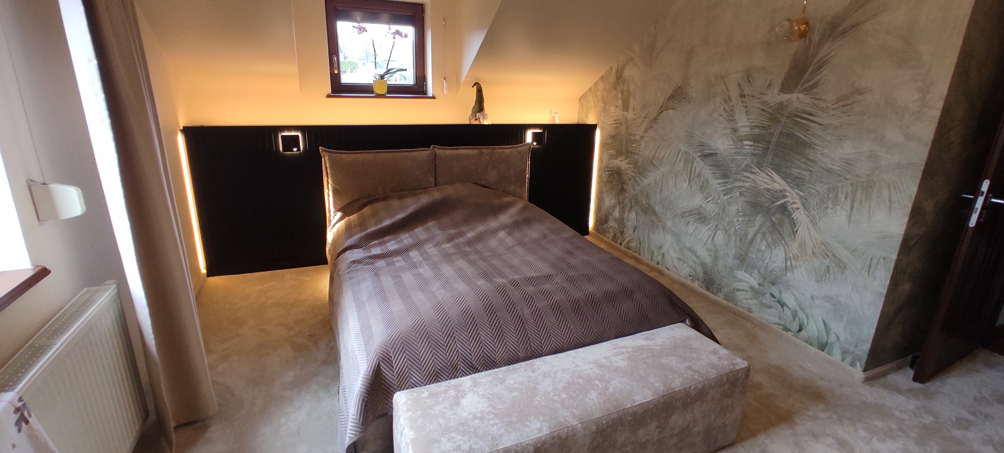 Sypialnia na poddaszu z ciemnym zagłówkiem z oświetleniem LED, wzorzystą tapetą z motywem tropikalnym i beżową tapicerowaną ławką.