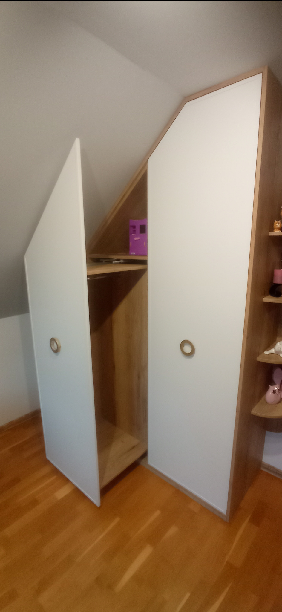 Szafa wnękowa na poddaszu z otwartymi drzwiami, widoczne półki i zabawki na półkach obok.