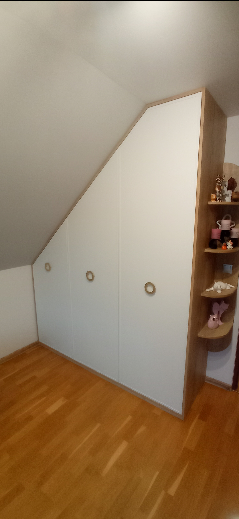 Biała szafa wnękowa z trzema drzwiami i okrągłymi uchwytami, dopasowana do skosu dachu, z drewnianymi półkami bocznymi, na których stoją ozdobne figurki, ustawiona na parkiecie.