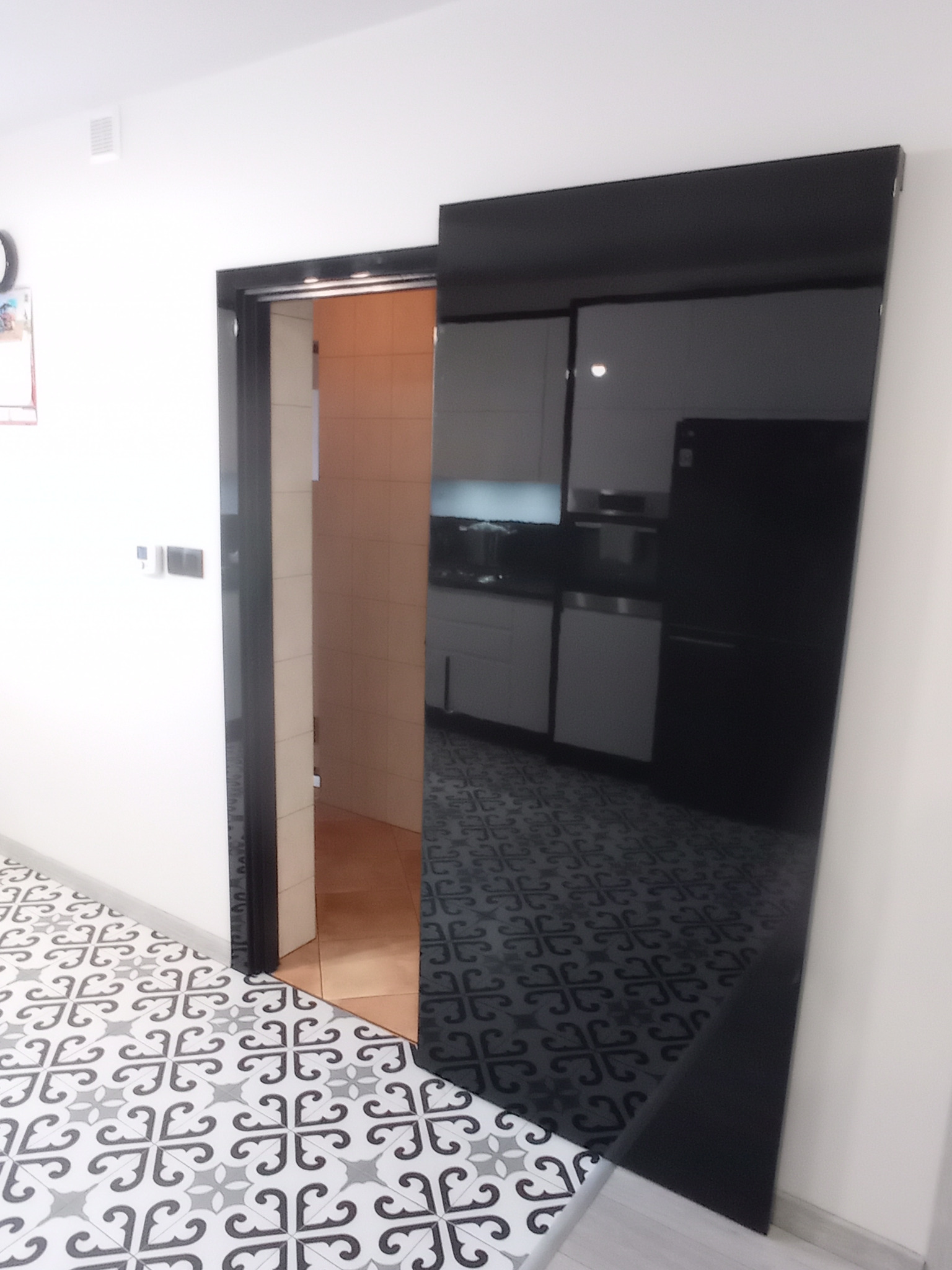 Czarne, przesuwne drzwi do kuchni z widocznym fragmentem białej zabudowy i lodówki, odbijające się w szklanej powierzchni drzwi, z wzorzystą podłogą w przedpokoju.