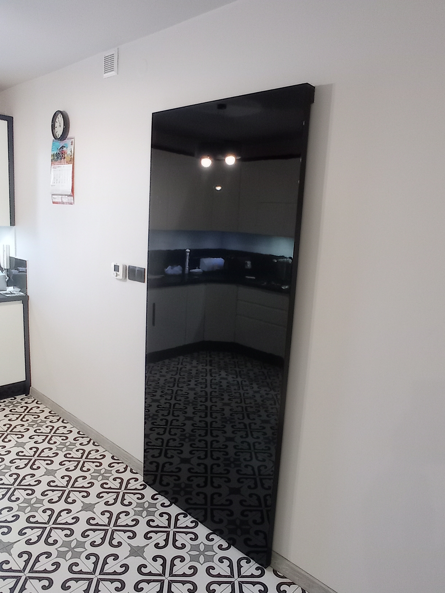 Wysoka, czarna szafa z lustrzanym frontem w korytarzu, odbijająca wnętrze kuchni z białymi szafkami i czarnym blatem, wzorzysta podłoga w biało-czarne ornamenty.