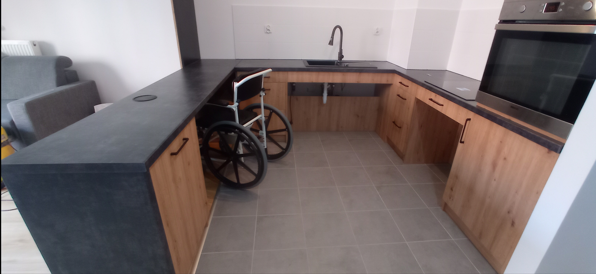 Kuchnia z blatem w kolorze grafitowym i drewnianymi szafkami, przystosowana dla osoby na wózku inwalidzkim; wózek stoi pod blatem, zapewniając przestrzeń do manewrowania.