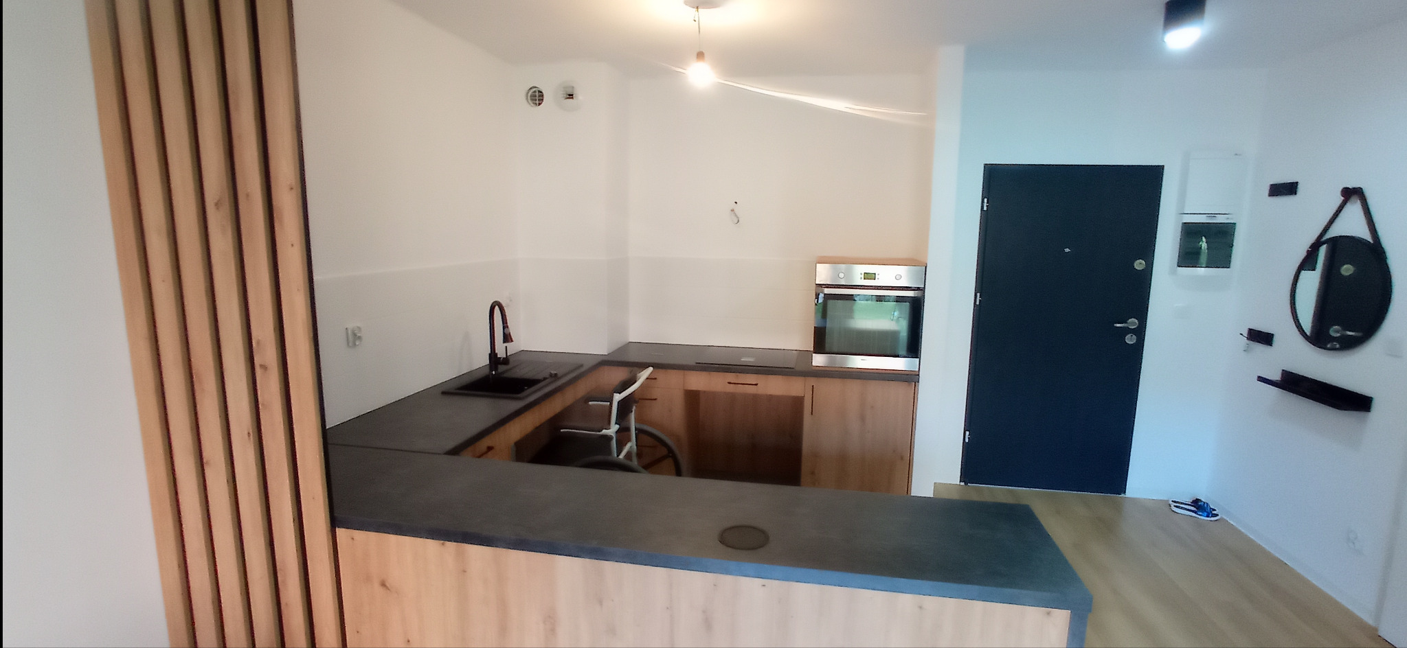 Nowoczesna kuchnia z blatem w odcieniach szarości i drewnianymi frontami, przystosowana dla osoby niepełnosprawnej poruszającej się na wózku inwalidzkim, widoczna czarna bateria i zlewozmywak.