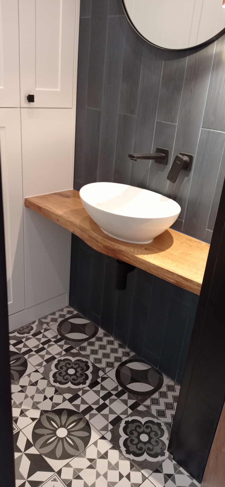 Biała, owalna umywalka nablatowa na blacie z jasnego drewna o nieregularnej krawędzi, z czarną armaturą i odpływem na tle ciemnoszarych płytek imitujących drewno i szafki z białymi frontami.