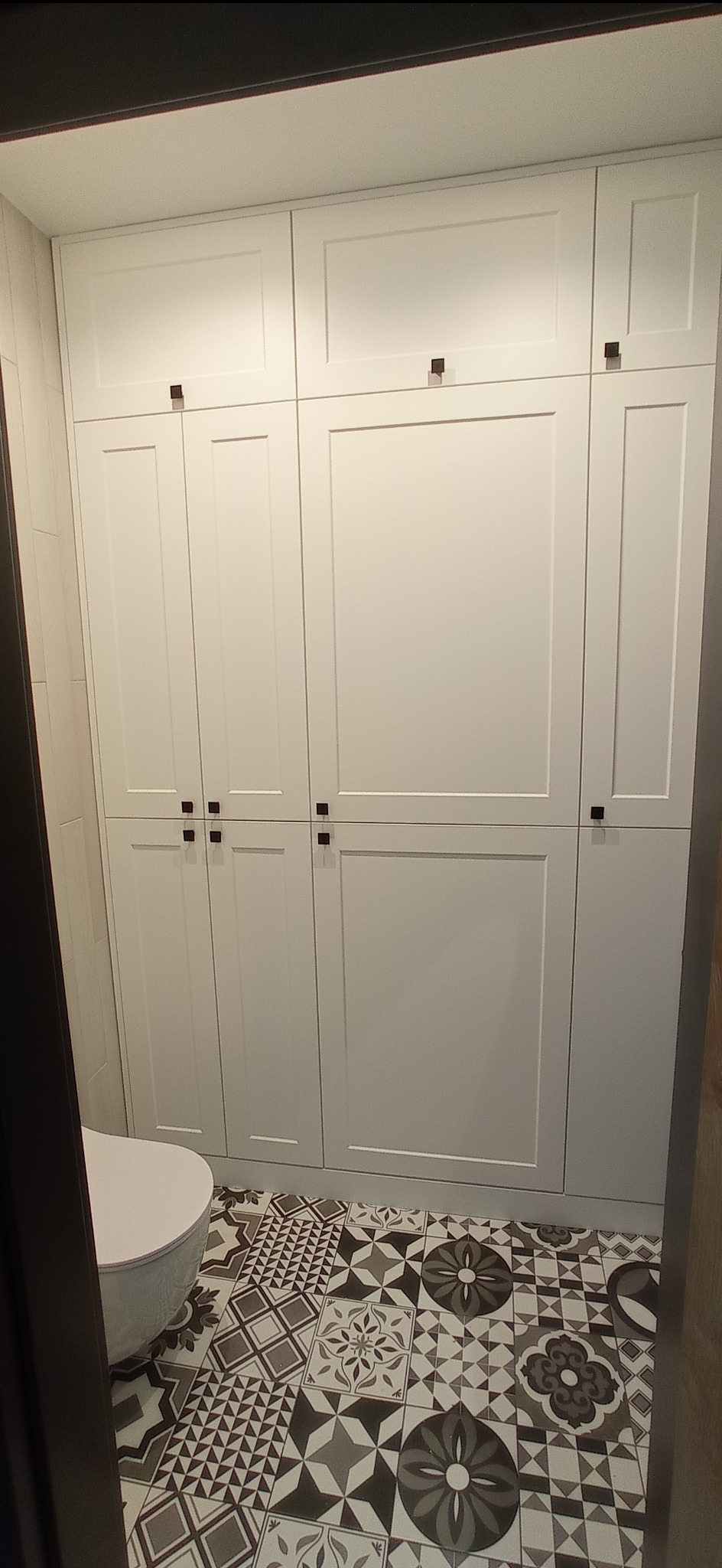 Biała szafa na wymiar z ciemnymi, kwadratowymi uchwytami w łazience z wzorzystą, czarno-białą podłogą i widoczną częścią wanny.