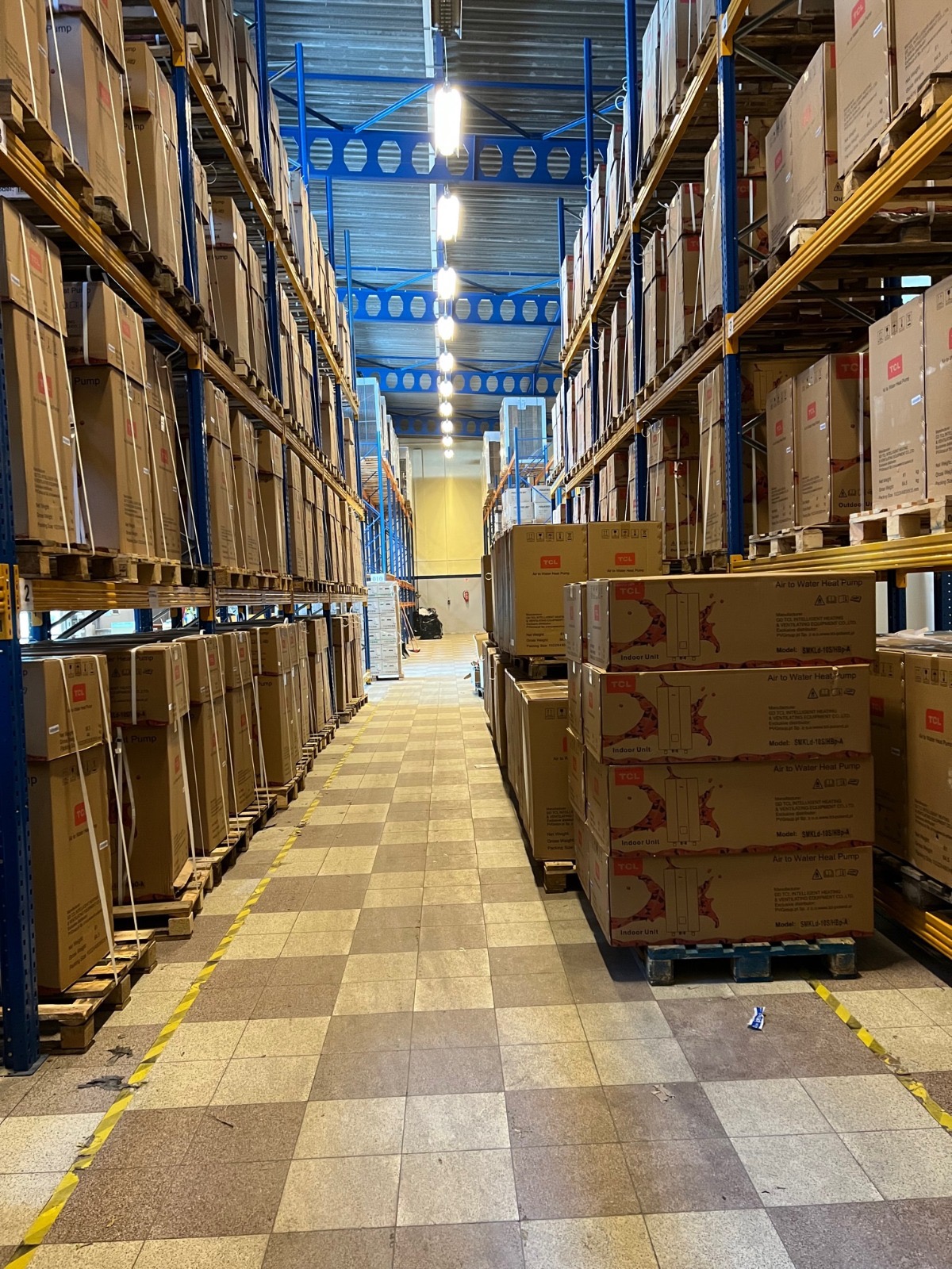 Magazyn z rzędami regałów wypełnionych kartonami, niektóre z oznaczeniami 'TCL Air to Water Heat Pump', widok w perspektywie wzdłuż korytarza, oświetlenie sufitowe.