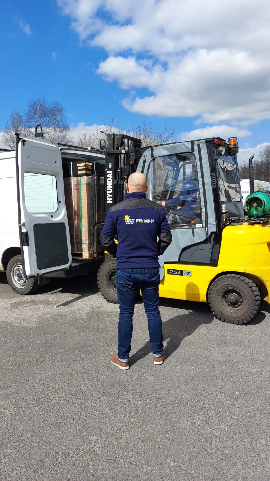 Pracownik w bluzie z logo PVgroup.pl nadzoruje rozładunek paneli z samochodu dostawczego za pomocą wózka widłowego Hyundai 25L-7.