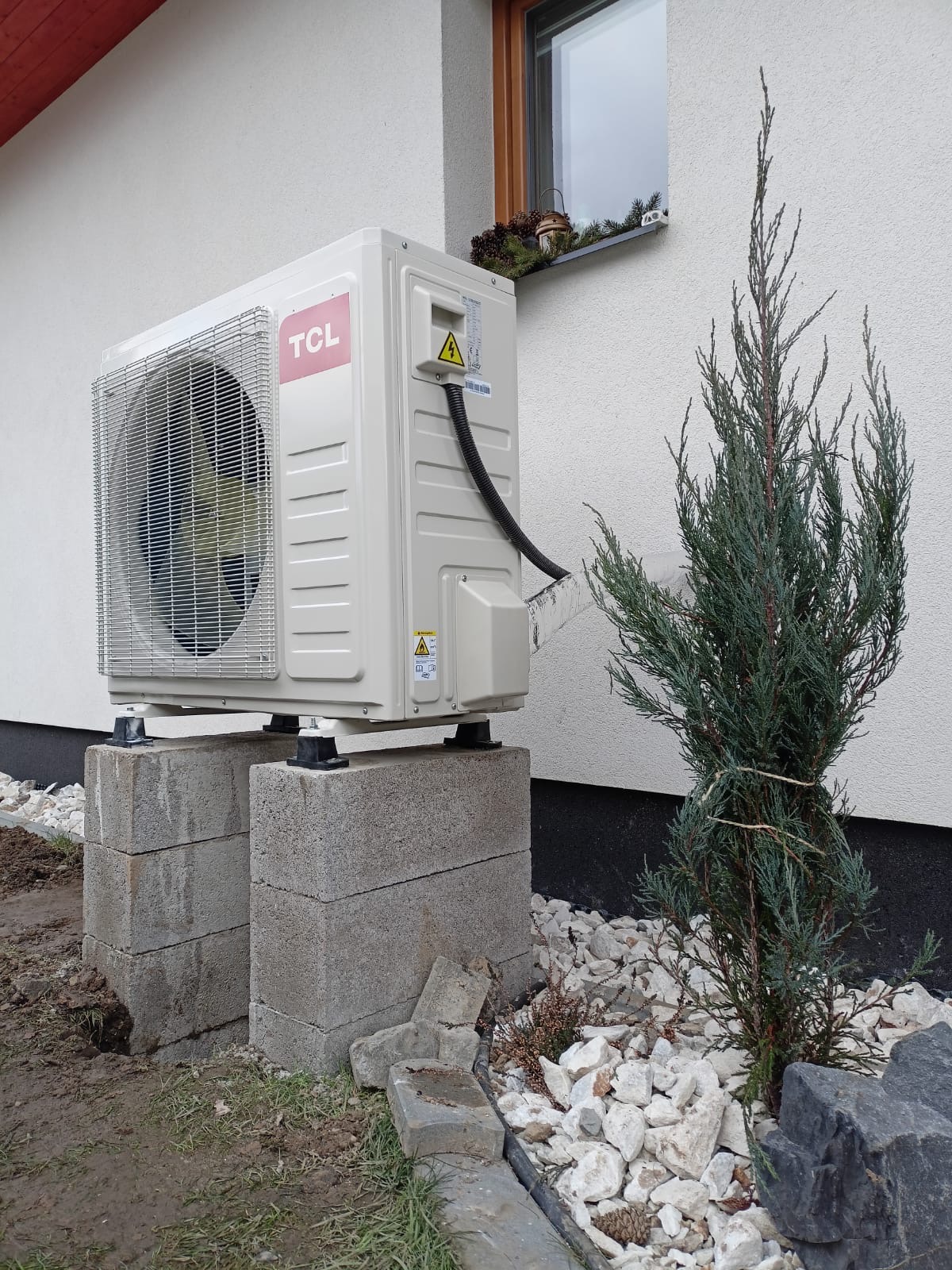 Biała jednostka zewnętrzna z logo TCL, zamontowana na betonowych bloczkach przy ścianie domu, obok iglastego krzewu i białych kamieni ozdobnych.