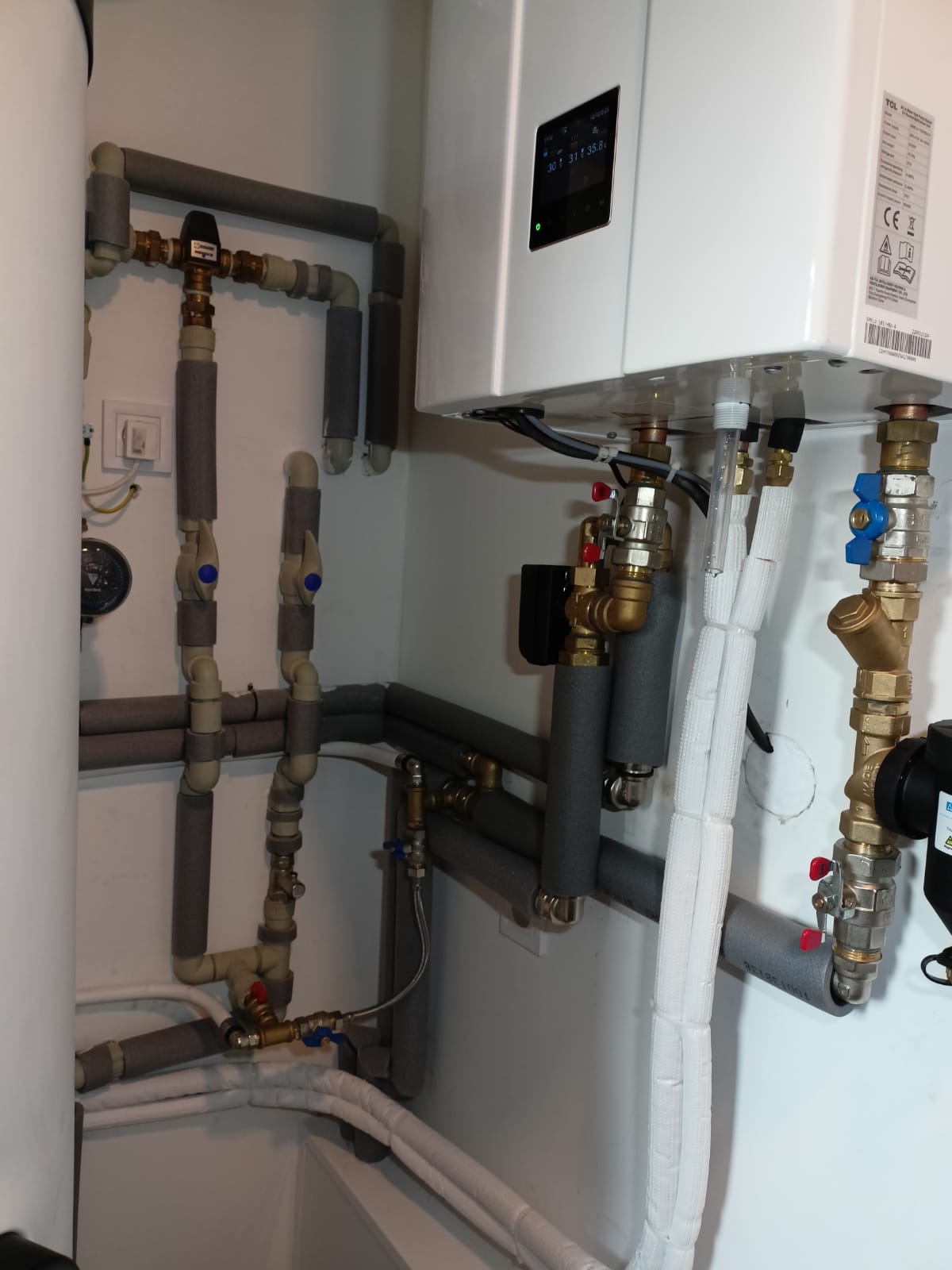 Biała instalacja centralnego ogrzewania z rurami w izolacji szarej i białej, zaworami z niebieskimi pokrętłami i wyświetlaczem temperatury.