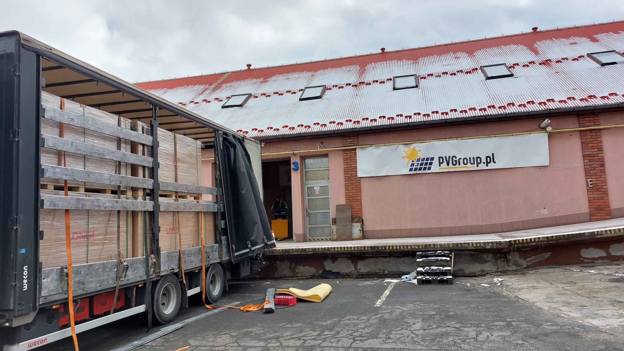 Ciężarówka z otwartą naczepą załadowana paletami z panelami słonecznymi na tle magazynu z banerem PVGroup.pl, widoczne elementy zabezpieczające ładunek.