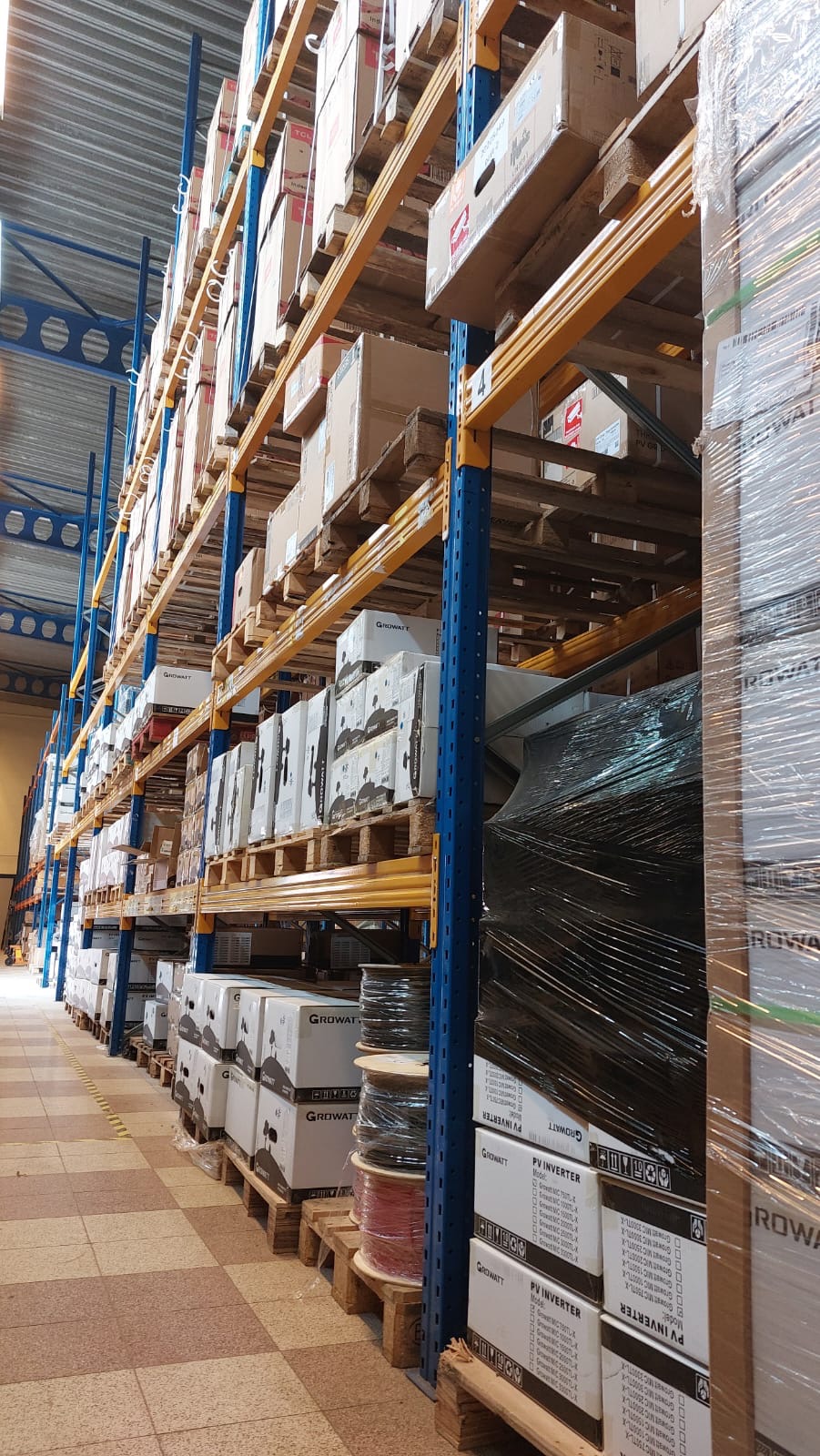 Magazyn z wysokimi regałami, na których ułożone są palety z kartonami, niektóre owinięte folią stretch, a także zwoje kabli. Na kartonach widoczne logo Growatt i oznaczenie PV Inverter.