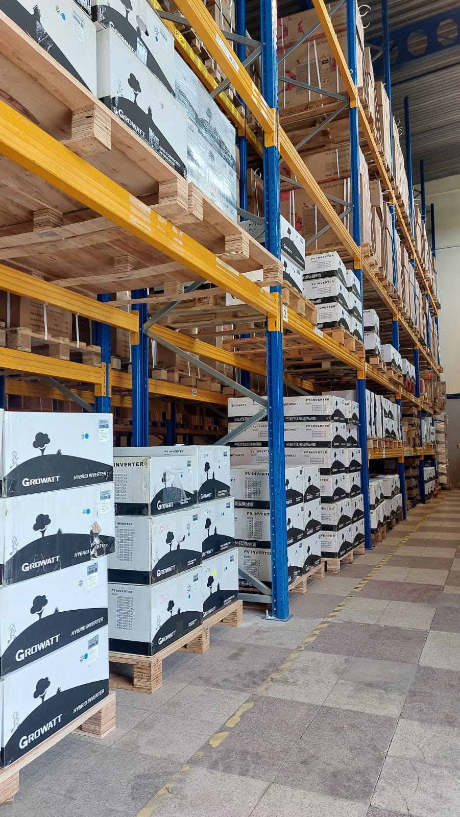 Magazyn z rzędami palet z kartonami falistymi zawierającymi inwertery fotowoltaiczne marki Growatt, widoczne niebieskie słupy i żółte belki regałów magazynowych.