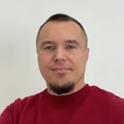 Portret mężczyzny z krótką fryzurą i brodą w czerwonej bluzie na białym tle.