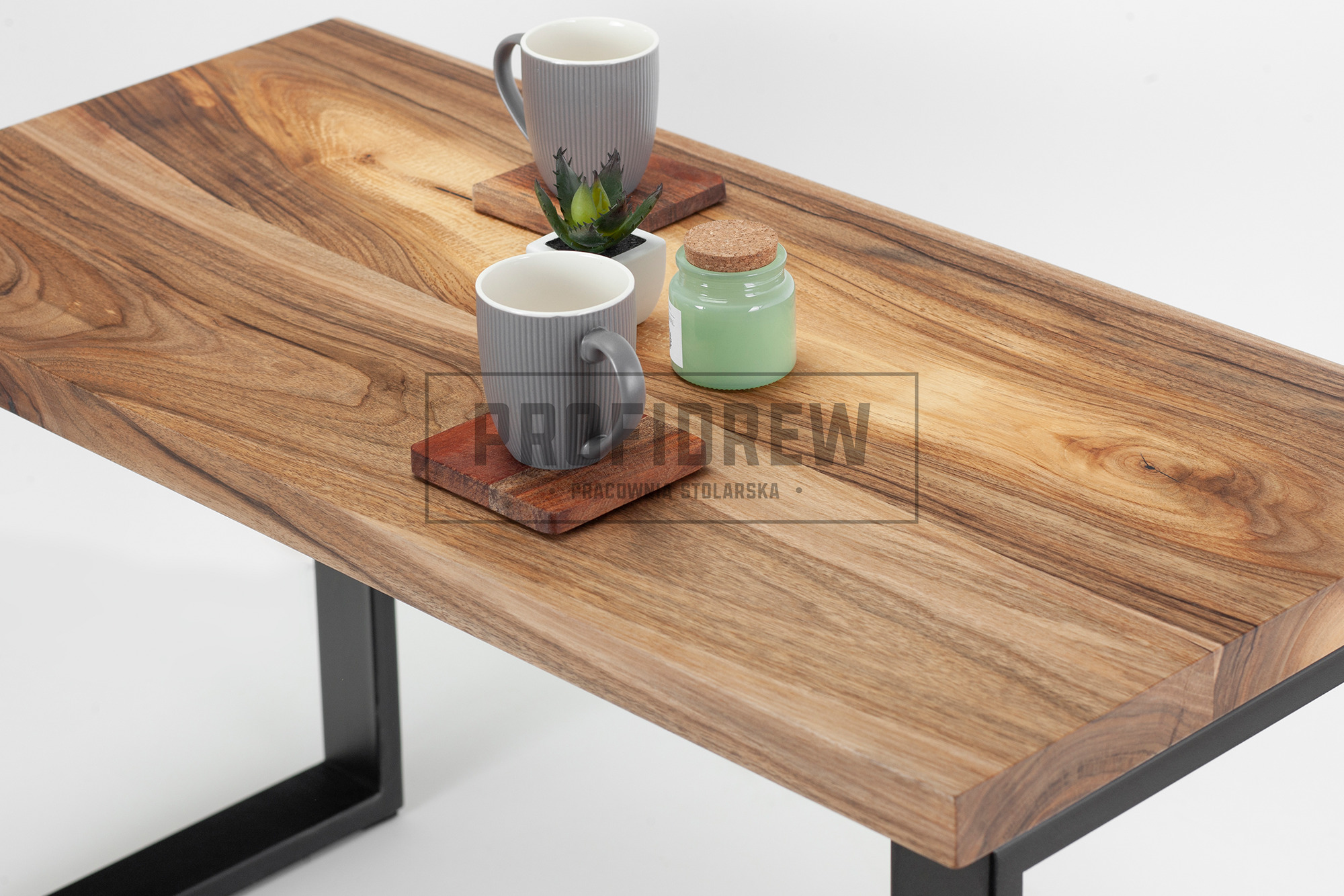 Stolik kawowy z blatem z litego drewna orzechowego, z widocznym usłojeniem, na czarnych, metalowych nogach. Na blacie dwie szare, prążkowane filiżanki na drewnianych podkładkach oraz mała roślina...