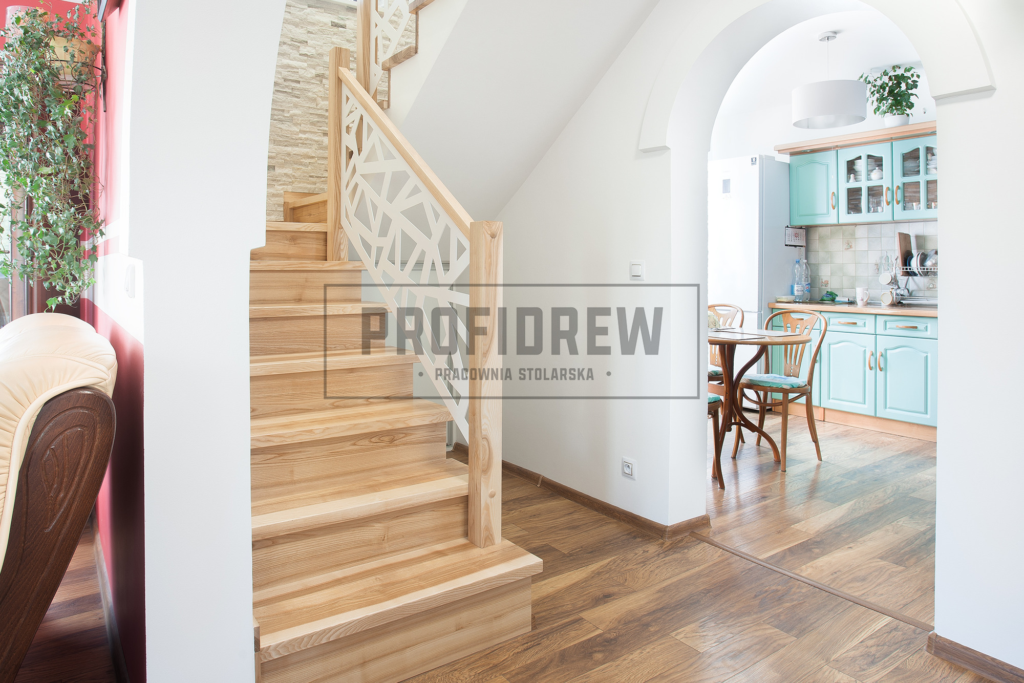 Jasne, drewniane schody z nowoczesną, białą balustradą o geometrycznym wzorze, prowadzące do jasnego wnętrza z widoczną kuchnią w pastelowych kolorach.