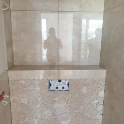 AgBud - Ściana w łazience wyłożona jasnymi, beżowymi płytkami ceramicznymi o zróżnicowanych wzorach i fakturach, z widoczną instalacją podtynkową do WC oraz odbiciem osoby robiącej zdjęcie w błyszczącej...