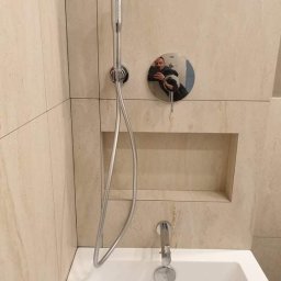 AgBud - Nowoczesna łazienka z beżowymi płytkami imitującymi marmur, chromowana bateria wannowa z prysznicem ręcznym, wbudowana półka w ścianie, odbicie fotografa w pokrętle termostatu.