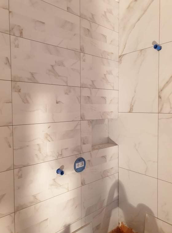 Ściana wyłożona białymi płytkami imitującymi marmur w łazience, widoczne niebieskie zaślepki na rury i wnęka w ścianie.