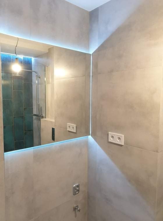 Wykończona łazienka z szarymi płytkami imitującymi beton, podświetlanym lustrem i widoczną kabiną prysznicową z turkusowymi płytkami odbijającą światło.