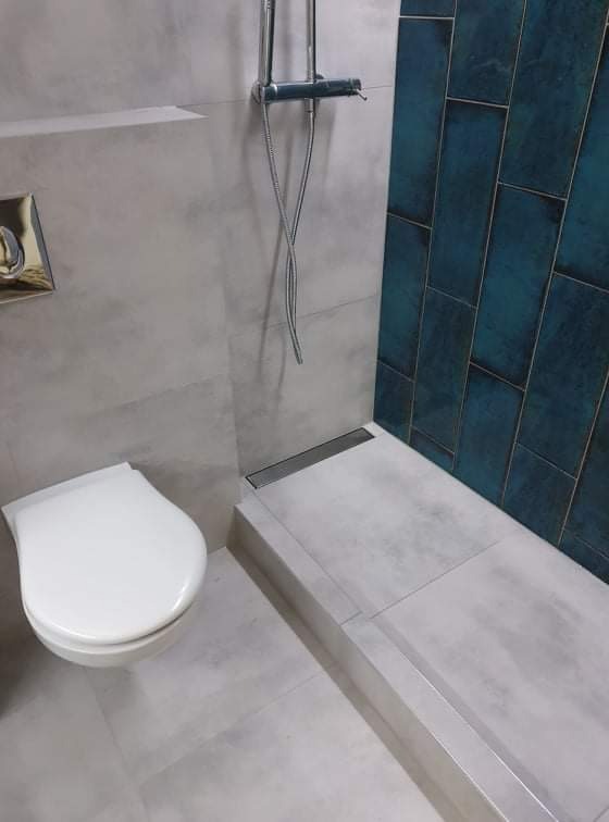 Nowoczesna łazienka z szarymi płytkami imitującymi beton, akcentem w postaci pionowych, turkusowych płytek na ścianie prysznica, minimalistycznym odpływem liniowym i podwieszaną toaletą.