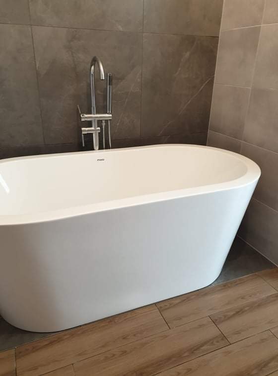 Wolnostojąca wanna z chromowaną baterią na tle szarych płytek i drewnopodobnej podłogi.