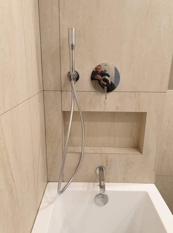 Nowoczesna łazienka z beżowymi płytkami imitującymi marmur, chromowana bateria wannowa z prysznicem ręcznym, wbudowana półka w ścianie, odbicie fotografa w pokrętle termostatu.