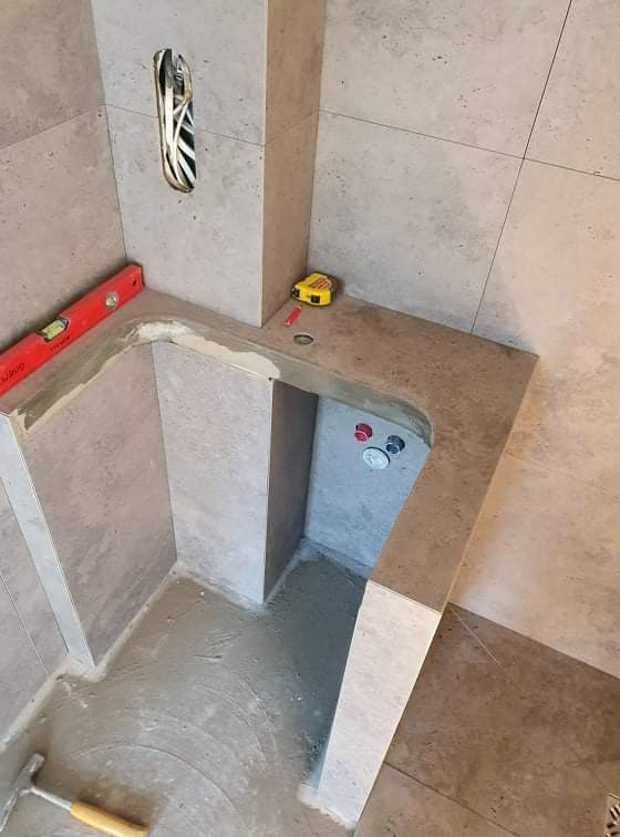 Narożnik w łazience w trakcie prac wykończeniowych, widoczne płytki w odcieniach szarości, poziomica, miarka zwijana, instalacja wodna oraz otwór z kablami elektrycznymi.