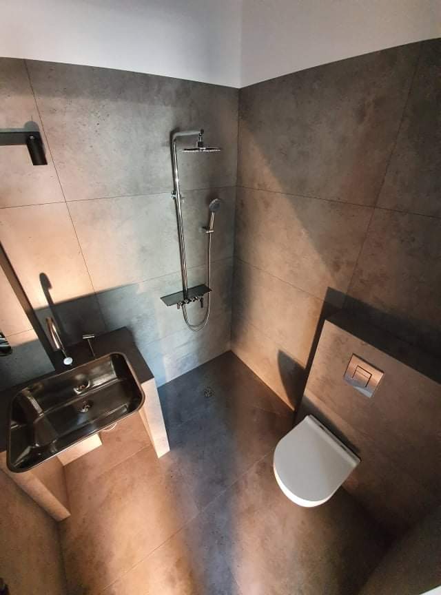 Nowoczesna łazienka w odcieniach szarości z metalową umywalką, chromowaną armaturą i podtynkową spłuczką, wykończona dużymi płytami ceramicznymi.