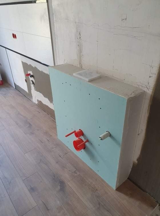 Stelaż podtynkowy zabudowany płytą gipsowo-kartonową z wyprowadzonymi rurami instalacyjnymi w łazience w trakcie prac wykończeniowych, widoczne drewnopodobne panele podłogowe i fragment ściany...