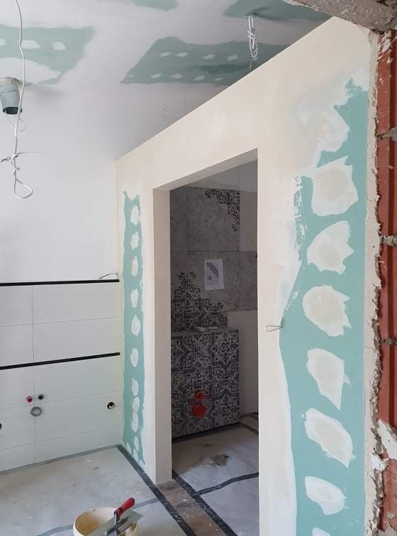 Łazienka w trakcie wykończenia: widoczne szpachlowane ściany, sufit z punktowymi łatami, przygotowane podłoże pod płytki oraz fragment ściany z cegły, zarys przyszłego wejścia do łazienki.