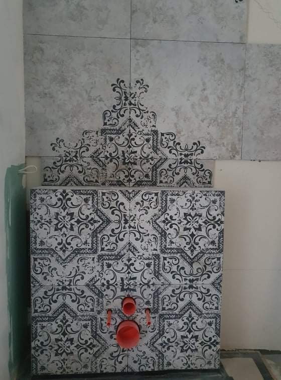 Ścianka w toalecie w trakcie wykańczania, obłożona dekoracyjnymi płytkami z motywem ornamentów, widoczne rury instalacji sanitarnej.