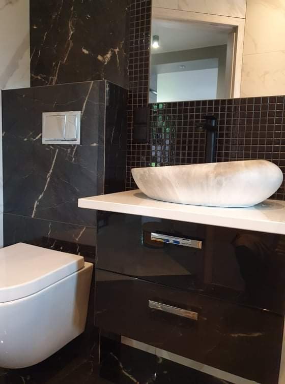 Nowoczesna łazienka z czarnymi marmurowymi płytkami, wiszącą toaletą i umywalką z jasnego kamienia na szafce z połyskiem, podkreślona przez czarną armaturę i mozaikę w tle.