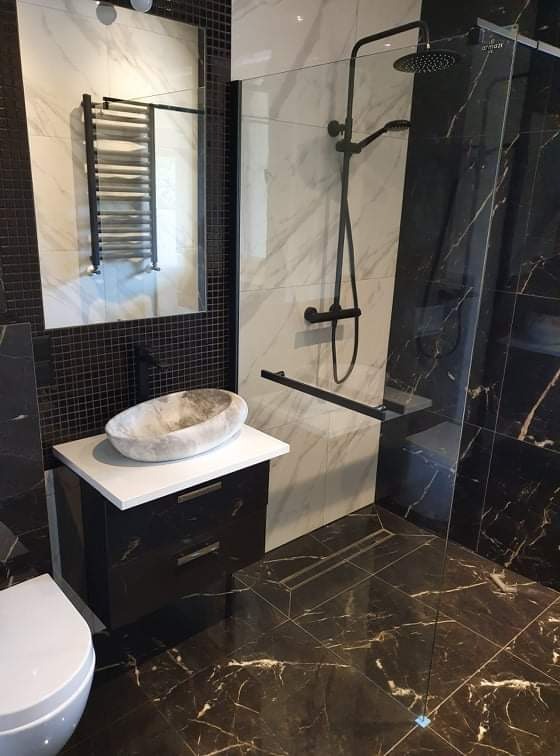 Nowoczesna łazienka z marmurową umywalką nablatową, czarną armaturą prysznicową i grzejnikiem drabinkowym odbijającym się w lustrze.