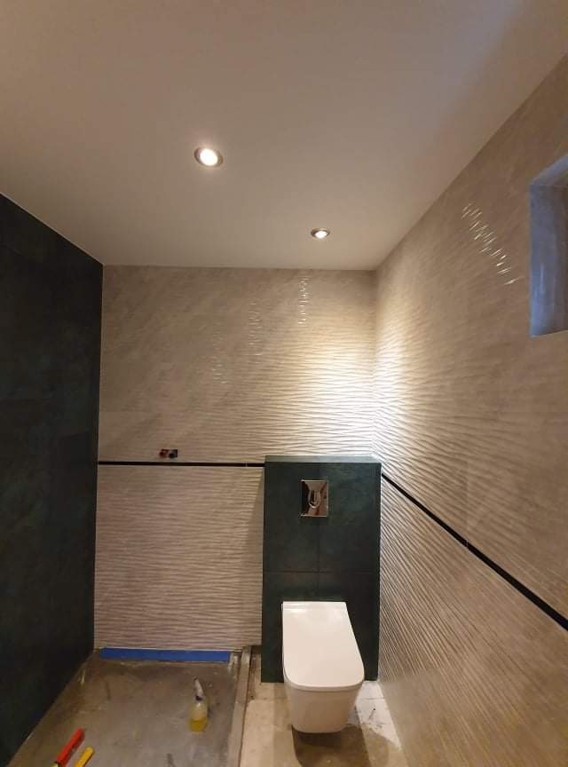 Nowoczesna łazienka w trakcie wykończenia, z podwieszanym WC na tle zielonej zabudowy i falistych, beżowych płytek ściennych z czarnymi akcentami. Na podłodze widoczne ślady prac glazurniczych...