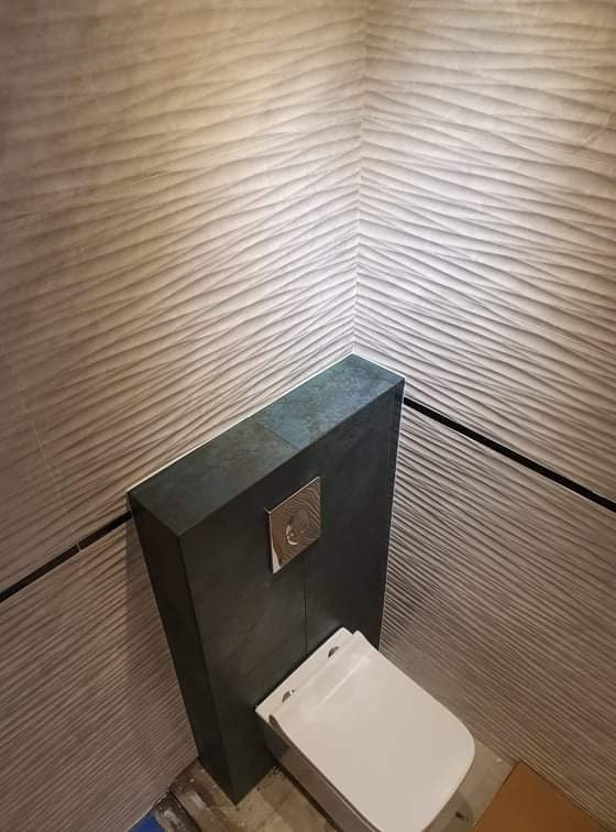 Nowoczesna toaleta podwieszana z szarym panelem i przyciskiem spłukującym na tle ściany wykończonej dekoracyjnymi płytkami o falistej strukturze.