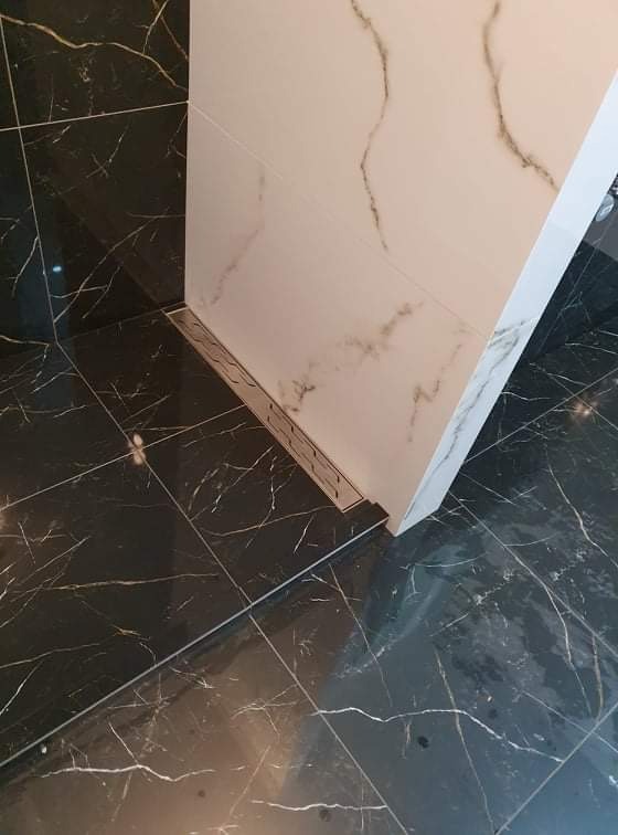 Detale wykończenia łazienki: czarne płytki podłogowe z białym żyłowaniem, białe płytki ścienne imitujące marmur i liniowy odpływ prysznicowy.