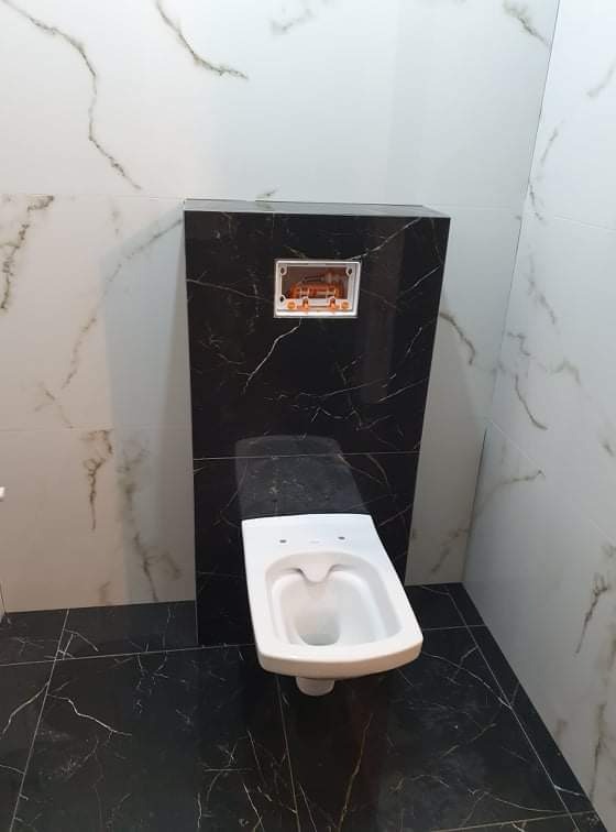 Wisząca miska WC w łazience wykończonej płytkami imitującymi marmur, widoczna puszka instalacyjna podtynkowego systemu spłukiwania na czarnym tle.