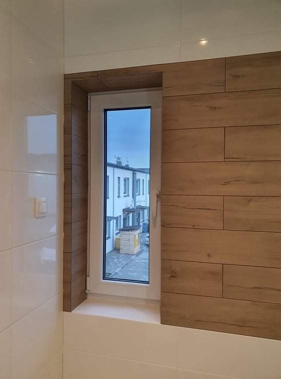 Wykończone wnęki okienne w łazience, z białymi płytkami po lewej i panelami imitującymi drewno po prawej stronie okna, widok na budynek z zewnątrz.