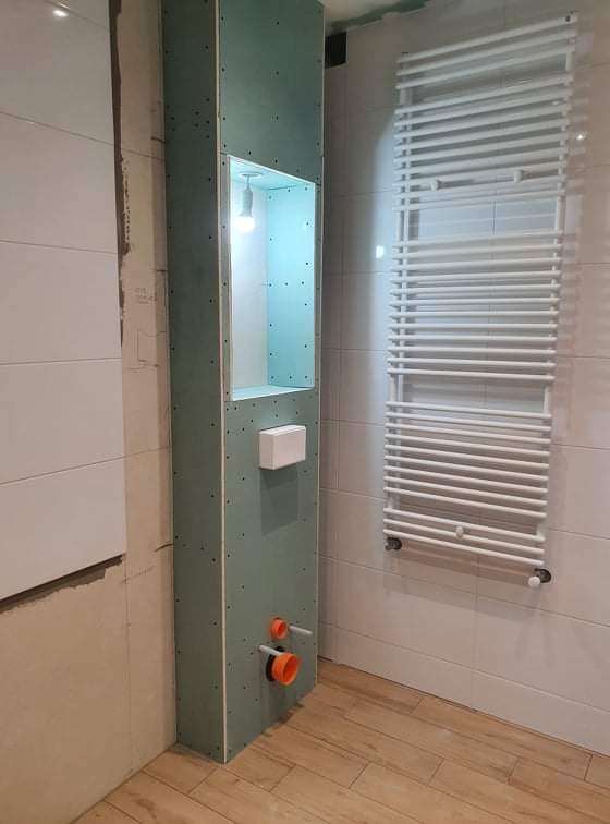 Łazienka w trakcie wykończenia: zabudowa z płyty gipsowej z wnęką oświetloną żarówką, widoczne rury instalacji wodnej i grzejnik drabinkowy na białych płytkach.