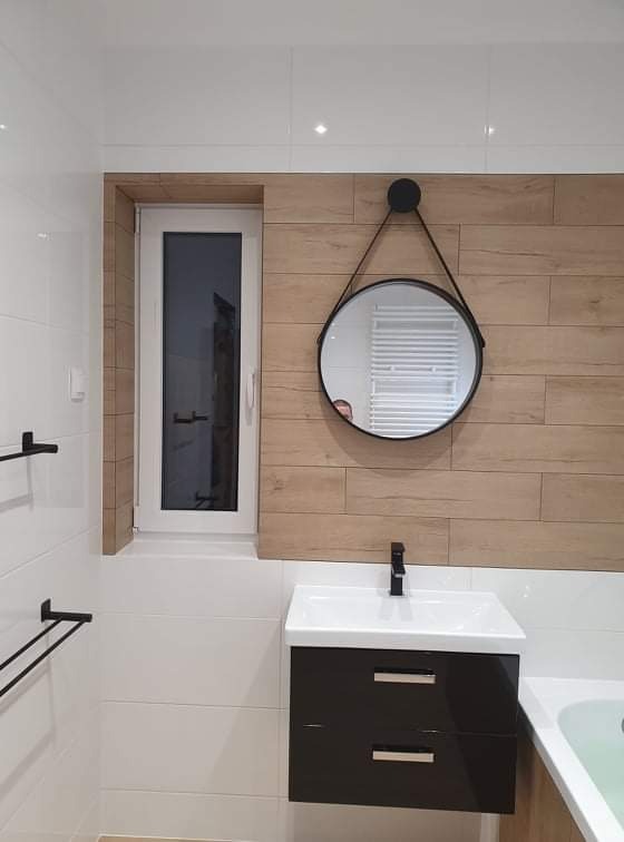 Nowoczesna łazienka z białymi płytkami, drewnopodobną ścianą, okrągłym lustrem na pasku i czarną szafką pod umywalką z czarną baterią.