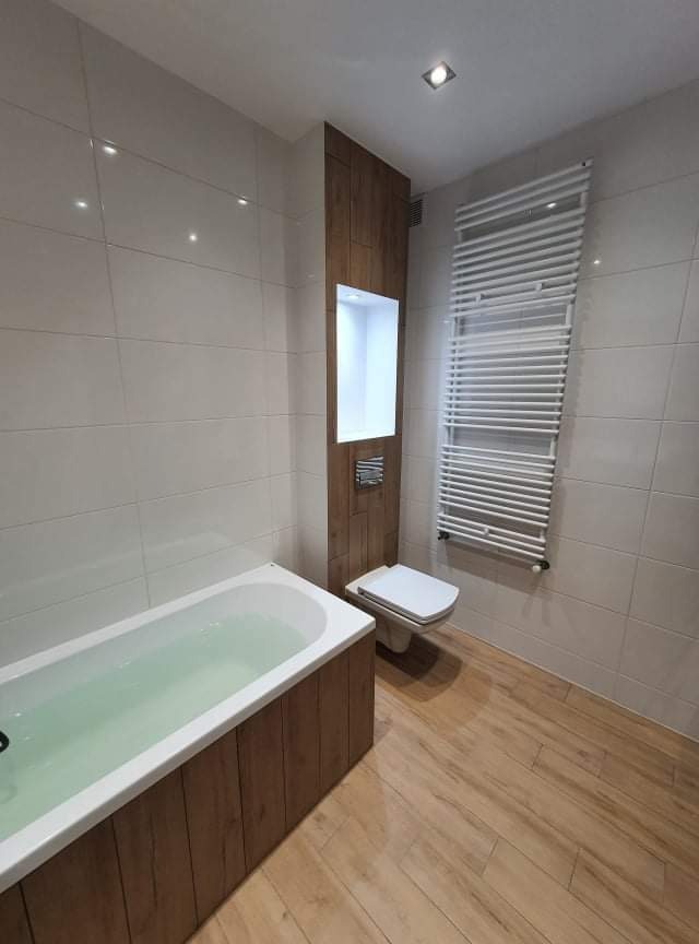 Nowoczesna łazienka z wanną wypełnioną wodą, toaletą, grzejnikiem drabinkowym i wnęką w obudowie z drewnopodobnych paneli, oświetlona górnym światłem.