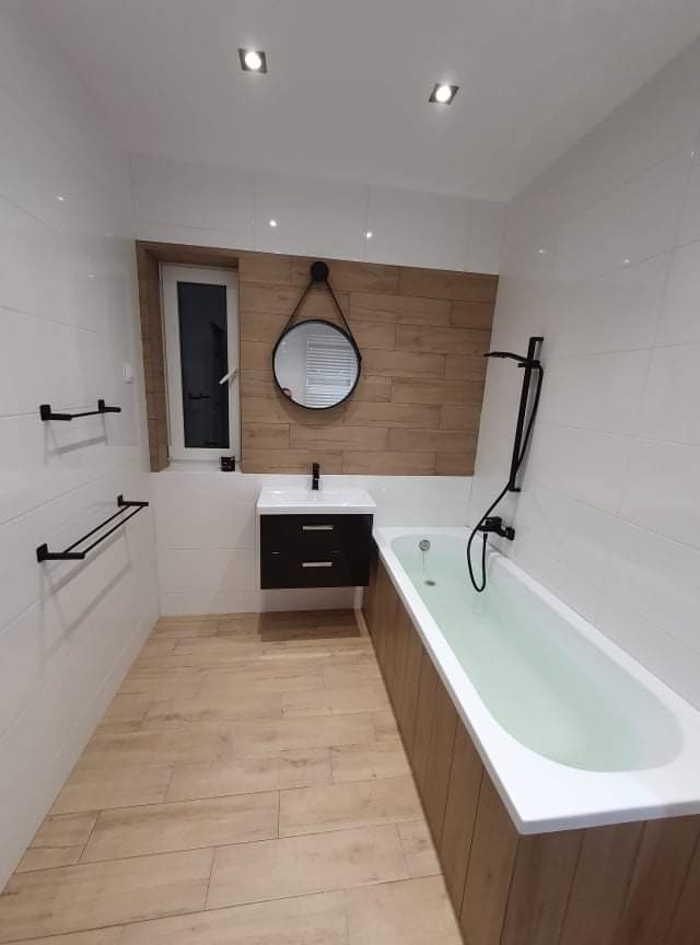 Wykończona łazienka w Zgierzu z białymi płytkami, drewnopodobną podłogą i ścianą, czarną armaturą i okrągłym lustrem.