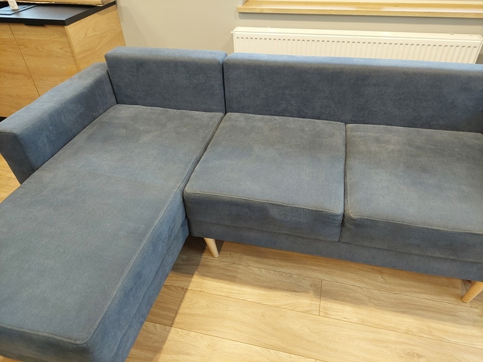 Szary narożnik z welurową tapicerką na drewnianych nóżkach, sfotografowany w Środzie Wielkopolskiej na tle jasnej podłogi i grzejnika.