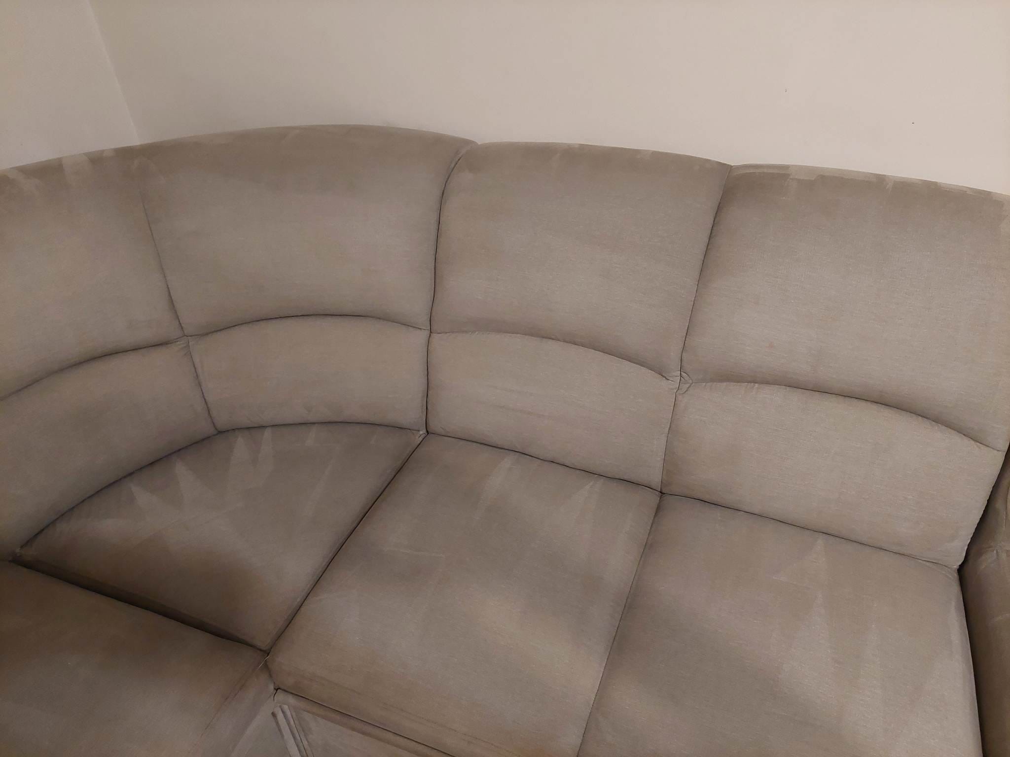 Fragment jasnoszarej, narożnej kanapy tapicerowanej z widocznymi śladami po czyszczeniu na poduszkach siedziska.