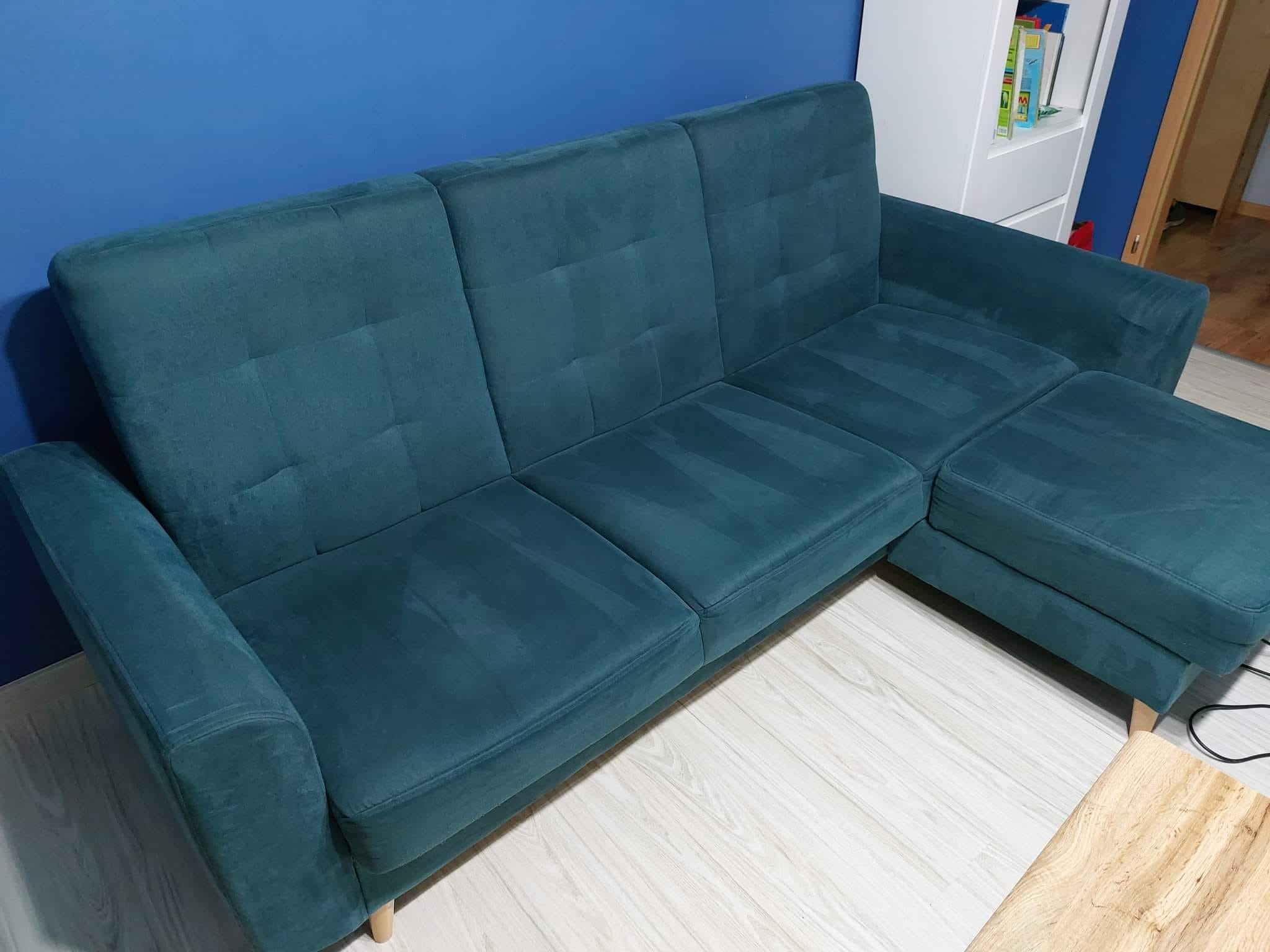 Turkusowa kanapa narożna z widocznymi śladami czyszczenia na siedzisku i oparciu, stojąca na jasnej podłodze, z niebieską ścianą w tle.