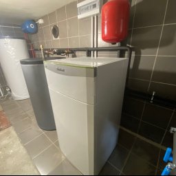 TB-INSTAL - Biały piec grzewczy Vaillant z szarym akcentem, obok szary zbiornik buforowy i biały zasobnik ciepłej wody. Czerwony zbiornik wyrównawczy na ścianie z szarymi płytkami. Widoczne rury instalacji...