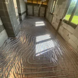 Białe rury systemu rozprowadzenia ciepła podłogowego ułożone na czerwonej siatce w surowym wnętrzu budynku w trakcie budowy, widoczne okna i ściany z betonu komórkowego.