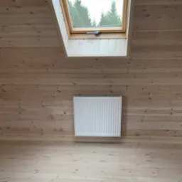 Biały grzejnik panelowy zamontowany na ścianie z jasnych, drewnianych desek pod oknem dachowym z widokiem na drzewa.