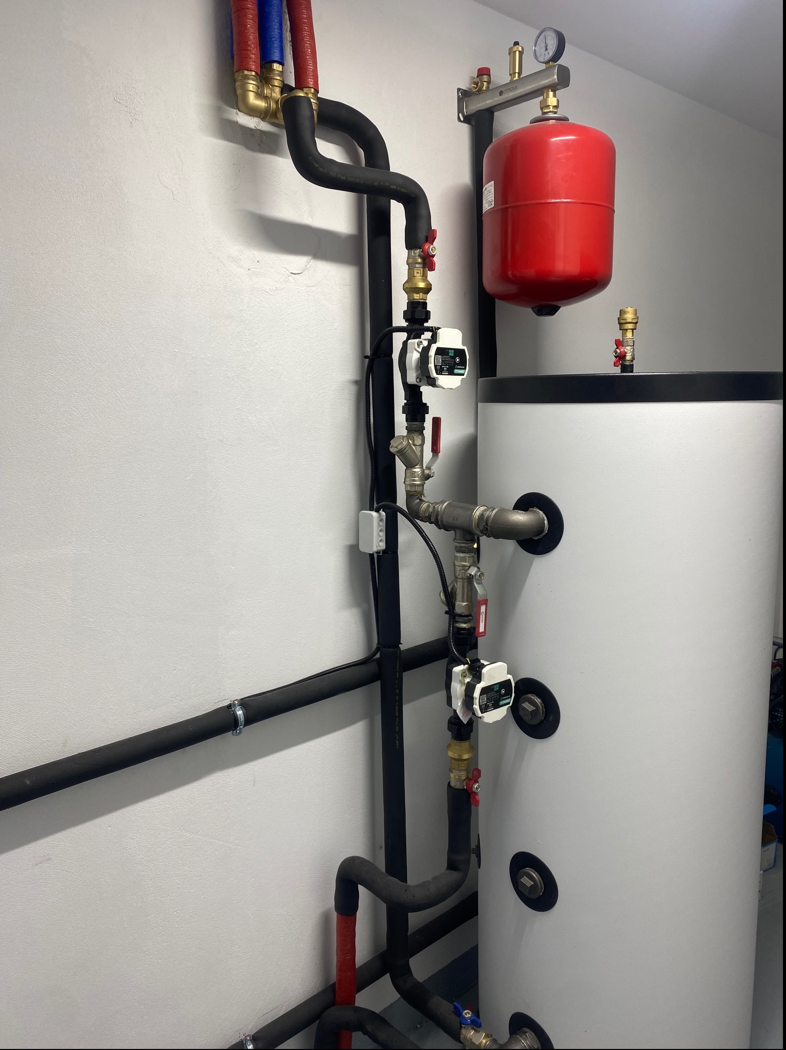 Biały zasobnik ciepłej wody użytkowej z podłączonymi rurami w izolacji, manometrem i pompami obiegowymi, zamontowany w pomieszczeniu technicznym.